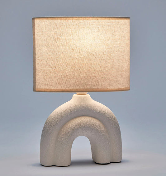 An illuminated white ceramic table lamp with a textured base and a white polyester shade, displayed against a plain blue background.
