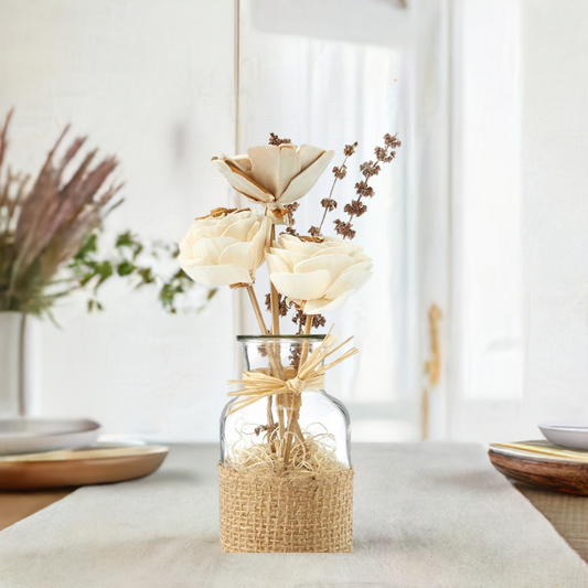 A clear glass jar containing an arrangement of natural dried flowers, tied with a rustic jute ribbon and adorned with bamboo sticks, cocoa fiber, shola flowers, wheat, palang, and teesi grass.