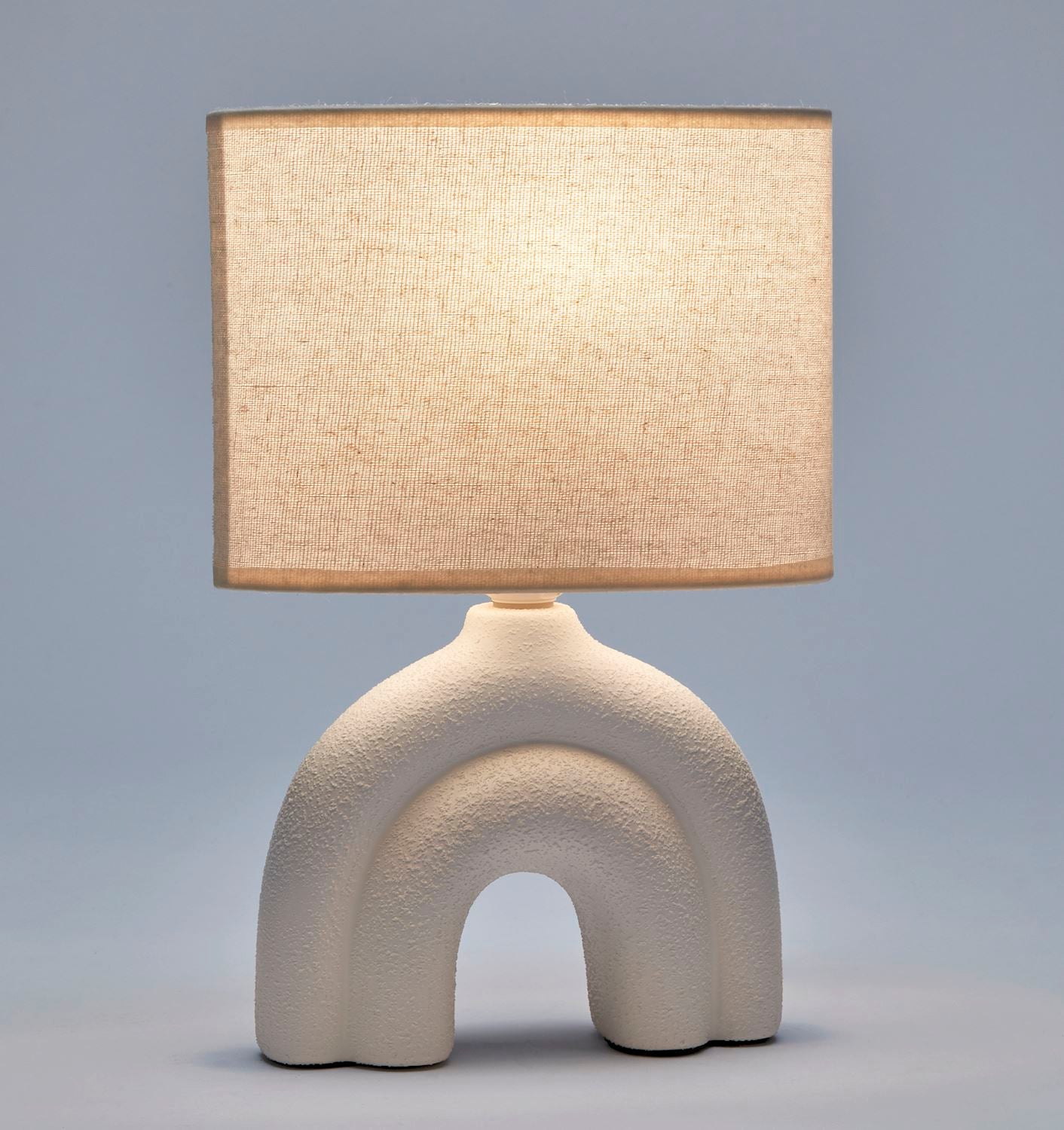An illuminated white ceramic table lamp with a textured base and a white polyester shade, displayed against a plain blue background.