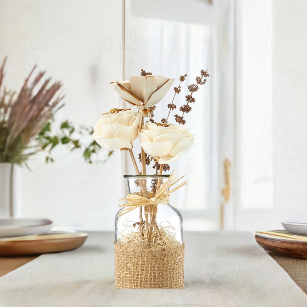 A clear glass jar containing an arrangement of natural dried flowers, tied with a rustic jute ribbon and adorned with bamboo sticks, cocoa fiber, shola flowers, wheat, palang, and teesi grass.